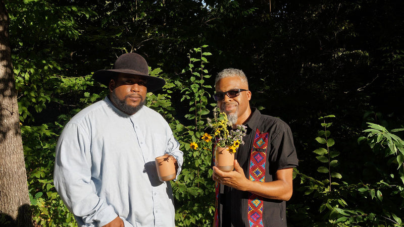 Two people are outside staring at the camera - the left person is wearing a button down shirt and hat and holding a mug, the other is wearing sunglasses and button down shirt, holding a mug that holds wildflowers