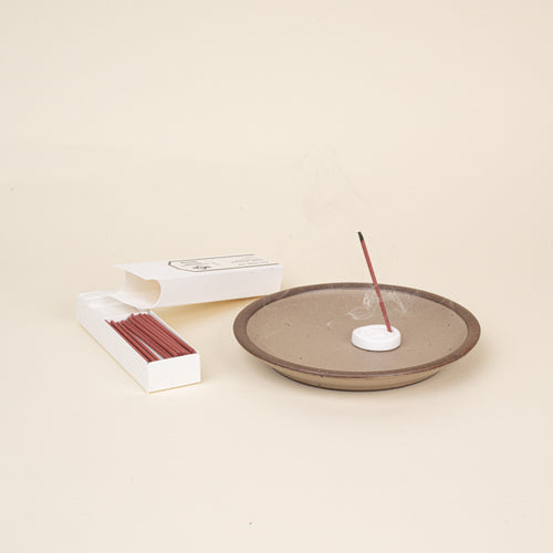 Lit stick incense in a holder on a ceramic dish, with open box of incense sticks beside it.