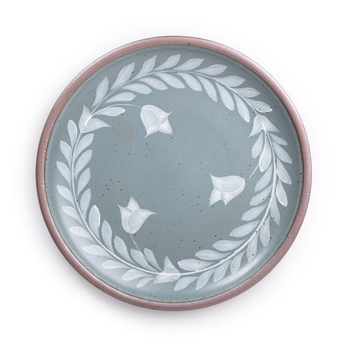 Large round ceramic platter with a hand-painted white leaf wreath and floral design on a blue-grey background with an exposed clay rim.