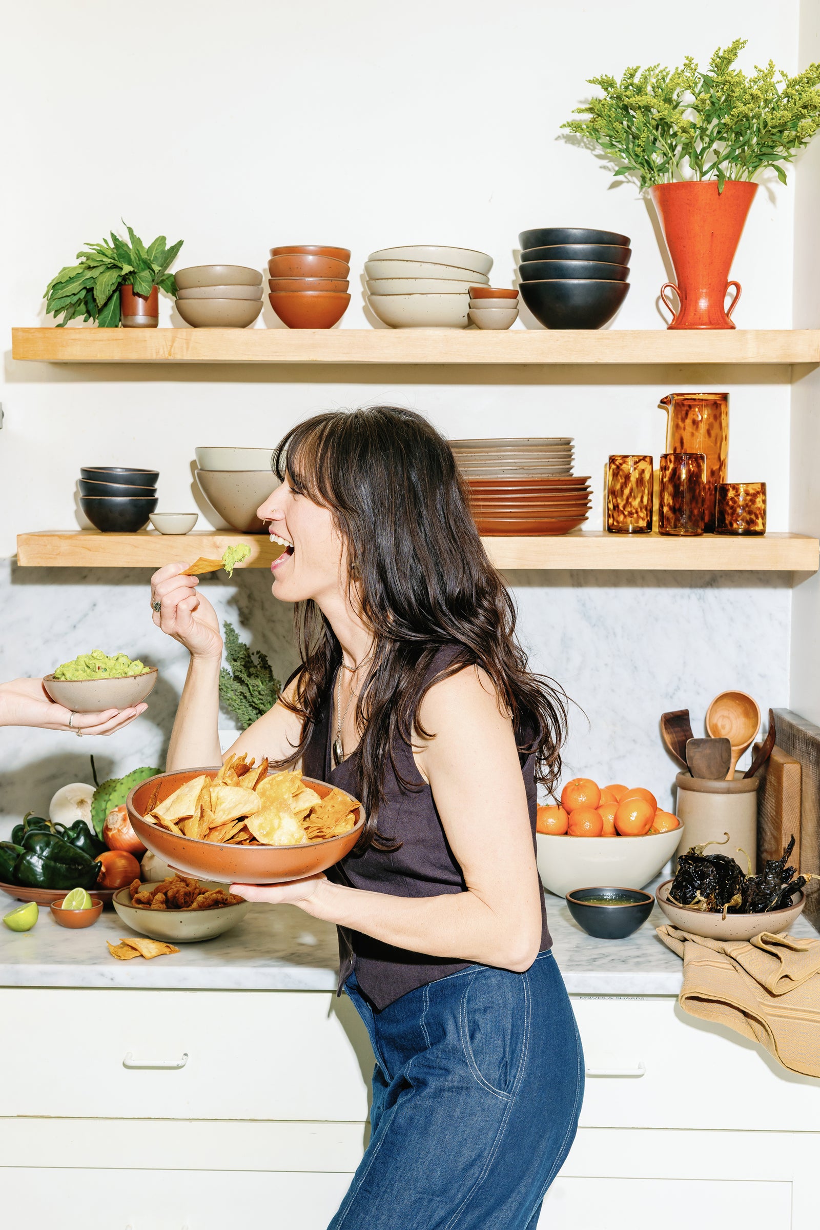 Connie in a kitchen holding a large shallow ceramic bowl filled with tortilla chips and someone holding a bowl of guacamole.